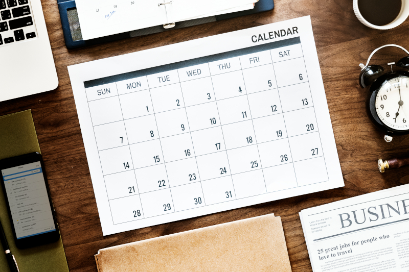 Calendar on desk surrounded by newspaper, laptop, clocks, organizer